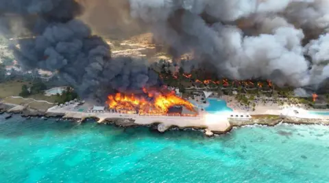 Reuters Aerial shot showing a large building on fire, and thick black smoke. The resort is on the beach and the water is vibrant blue.