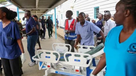 Ministry of Health Kenya A person wearing a blue t-shirt pulls a gurney, his mouth open as he looks at the camera and appears to move quickly. Other first responders wearing scrubs or high-vis vests or uniforms stand around, appearing to look forwards in the same direction at something out of view.