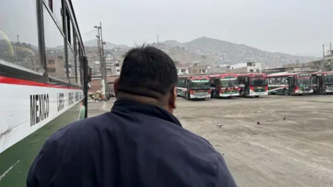 BBC Bus driver Toño photographed from behind standing next to a bus in a large parking area