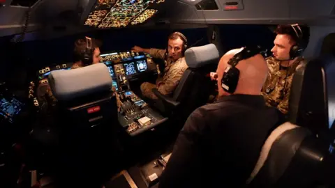 Lee Durant/BBC BBC correspondent Jonathan Beale is seen wearing a black shirt and headset in the cockpit of the Voyager. Two pilots, another crew member and the large control system can be seen in the background