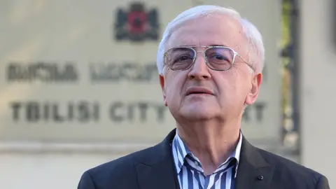 Reuters Miss Culley's lawyer, Malkhaz Salakaia, is standing in front of Tbilisi City Court in Georgia, though the sign on the wall behind him saying this is out of focus. He is wearing a black suit jacket and an open-necked shirt with thick stripes in dark blue and white. He has silver rimmed glasses and short, white hair and has his head held confidently upright, chin slightly jutting out.