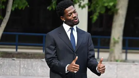 Getty Images Hill flashed thumbs up to reporters after the hearing. Nas X wearing a suit and with his thumbs up and smiling