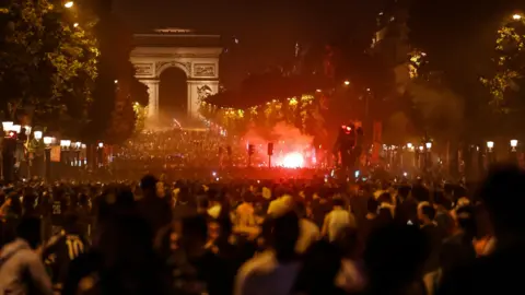 AFP via Getty Images Bustling crowds swamp Avenue des Champs-Élysées, lit up by a ball of red flares along the landmark mall. The Arc de Triomphe can be seen at the end of the avenue.