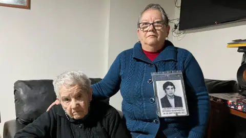 Grace Livingstone Ana Aguayo's mother, wearing a black cardigan and sitting in a leather chair, looks into the camera as her daughter has her hand placed on her shoulder. Ana has the photo of her brother pinner to her cardigan. 