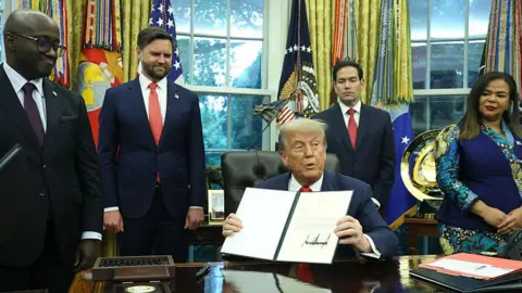 Getty Images Seated in the White House’s Oval Office, Donald Trump holds a letter of congratulations. Standing around the US president are Rwanda Minister of Foreign Affairs and Cooperation Olivier Nduhungirehe, US Vice-President JD Vance and Secretary of State Marco Rubio, all in dark blue suits, white shirts and red or, in the case of Nduhungirehe, blue, ties, and Democratic Republic of the Congo Foreign Minister Thérèse Kayikwamba Wagner, in a blue patterned shirt and skirt.
