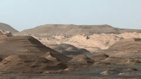 NASA The picture shows the higher regions of Mount Sharp on Mars. In the foreground are some darker areas with some small hillocks with a slightly larger slope off to the left. In the middle of the photo are some lighter more craggy looking rocky areas. In the distance are bigger hills that are more rounded with gentle slopes. The sky, which is visible at the very top is a dark grey.