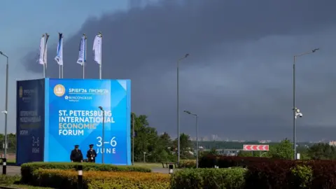 Reuters Police officers stand in front of a banner and flags with the logo of the St. Petersburg International Economic Forum (SPIEF) as heavy smoke billows in the background after Ukrainian drones hit infrastructure