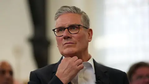 Getty Images Sir Keir Starmer, with short grey hair and black glasses, stands up while wearing a white shirt with a black suit jacket. The background behind him is blurred.
