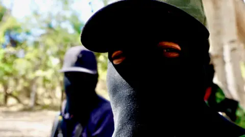 Darren Conway/BBC A cartel member wearing a black balaclava looks into the camera, another - also wearing a balaclava, stands in the background.