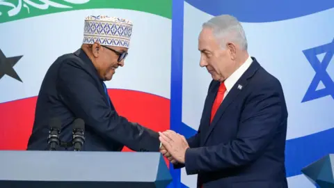 Prime Minister of Israel's Office Abdirahman Mohamed Abdullahi and Benjamin Netanyahu shake hands. In the backdrop are the flags of Somaliland and Israel.