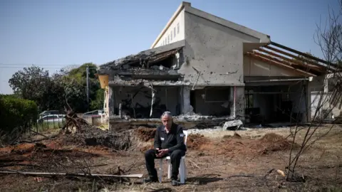 Reuters A man sits outside a house in central Israel that was damaged by a projectile launched by Hezbollah in Lebanon (12 March 2026)