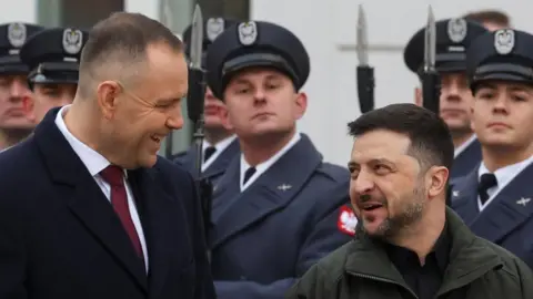 Reuters Polish President Karol Nawrocki and Ukrainian President Volodymyr Zelensky smile at each other while soldiers stand to attention behind them.