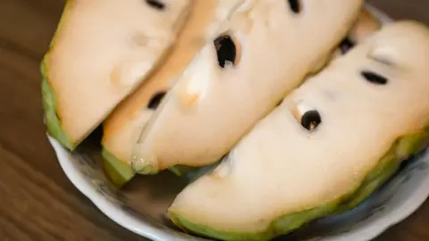 Getty Images Close-up of the white flesh, black seeds and green skin of a sliced atemoya on a plate.