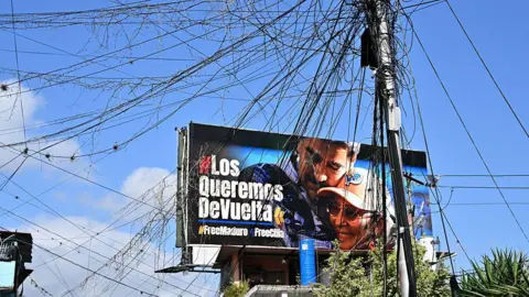 Juan BARRETO / AFP via Getty Images A spider web of power lines can be seen in front of a billboard calling for the releas of ousted Venezuelan President Nicolas Maduro and his wife, Cilia Flores, inthe Petare neighborhood, Caracas. 