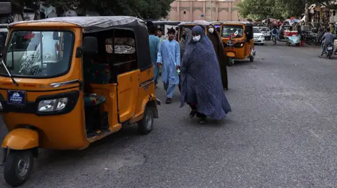 AFP via Getty Images A woman covered with a long robe from head to toe, with her face covered by a medical mask, walks down the street near a tuktuk. Men can be seen in the background