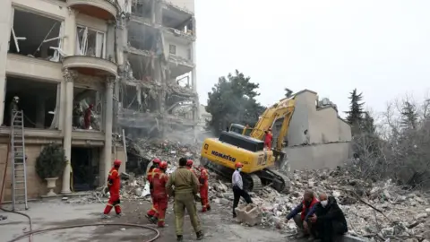 EPA Emergency workers wearing red and grey outfits at the site of a residential building damaged in an attack where a yellow digger is in the rubble; three people wearing civilian clothes, one wearing a red helmet and two face masks, are also in the picture, two of them sitting on the curb edge.