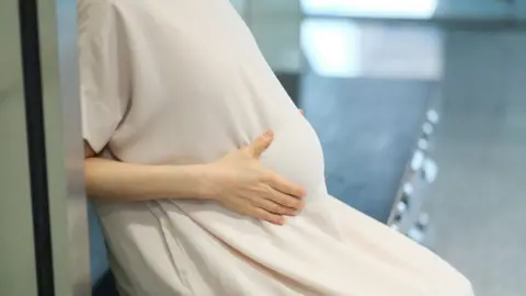 Getty Images Close-up of a pregnant woman's torso, with her hands cupping her baby bump. She is wearing a long beige dress and sitting on a bench.