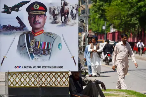AFP via Getty Images A man sits beside a poster showing a Pakistani military officer in full ceremonial uniform, decorated with medals and insignia, set against images of fighter jets, missiles and galloping horses.