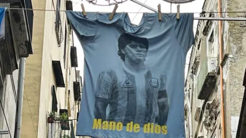Laura Lezza/Getty Images) A T-shirt with the face of Maradona and the inscription 'Mano de dios' on the streets of the historic centre the day after the first anniversary of the death of Diego Armando Maradona on November 26, 2021 in Naples, Italy. 
