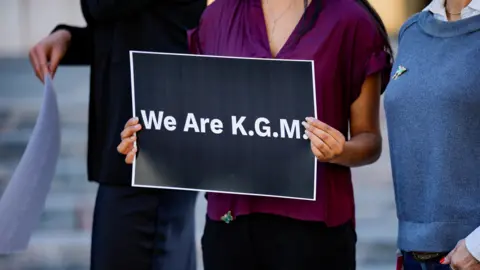 Reuters A woman wearing a purple shirt holds up a sign that reads: We are K.G.M.