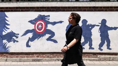 AFP via Getty Images A woman wearing a black dress walks past an anti-US mural outside the former US embassy in Tehran on 15 June. 