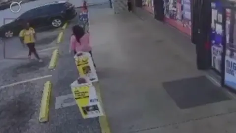 @realDonaldTrump / via Truth Social An image from surveillance video shows a woman in a pink blouse standing outside a convenience store as a man in black pants and a yello shirt approaches her from the left