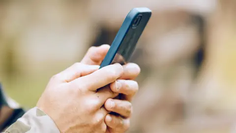 Getty Images A pair of hands holding a mobile phone, with a camouflage cuff showing. This is a stock image showing a model.