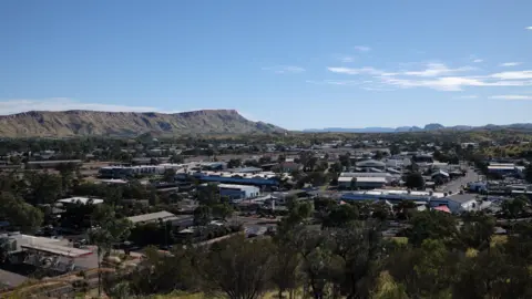A Panoramic view of the town of Alice Springs where you can see mountains, some houses and warehouses.