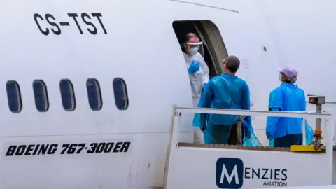 Reuters Man in full PPE talks to man and woman wearing blue plastic outer clothing and face masks at the entrance to a Menzies Aviation Boeing 767 plane.