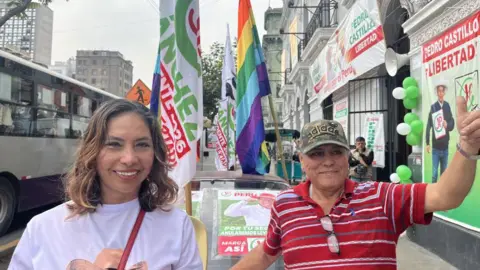 María is wearing a white Sánchez T-shirt, standing in the street next to campaign flags and a man in a red polo shirt