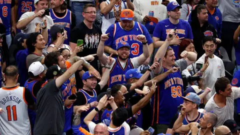 Getty Images Rows of people mostly wearing jerseys in the Knicks colors of orange and blue throw their hands in the air, cheering a victory.