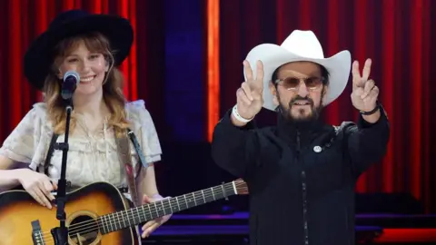 Getty Images Molly Tuttle and Ringo Starr performed at The Grand Ole Opry in Nashville, Tennessee last year. Molly is wearing a cream-coloured blouse and black hat while holding a guitar. Star has on a white cowboy hat and a black jacket and is doing the peace sign.