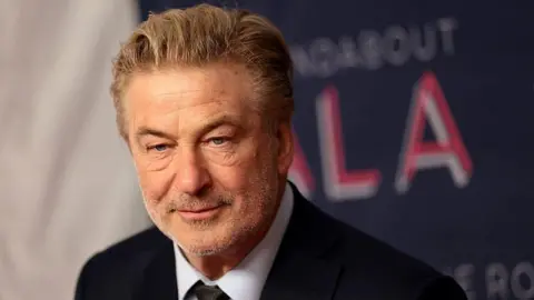 Getty Images Alec Baldwin looks ahead in a suit and tie. His greying hair is slicked back and he is wearing a soft smile. In the background is a red carpet backdrop in which the word 