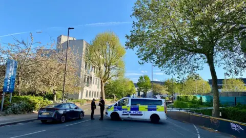 BBC Police tape closes off the road. Two officers can be seen stood next to a police vehicle. The area is leafy with a pale building in the background. 