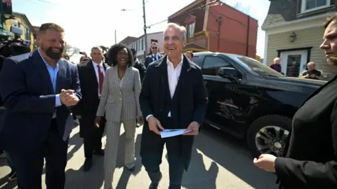 Reuters Mark Carney, in a dark suit and no tie, smiles as he walks outside with Terrebonne candidate Tatiana Auguste, in a grey pinstripe suit, on a sunny day.