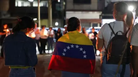 MIGUEL GUTIÉRREZ/EPA/Shutterstock Relatives of political prisoners hold candles and posters during a vigil in Zamora, Venezuela, 26 February 2026. A woman, with her back to the camera, has a Venezuelan flag wrapped around her shoulders. Others hold candles.