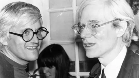Getty Images David Hockney and Andy Warhol in the 1970s. They look like mirror images with their white hair and round glasses.