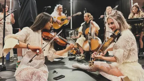 Kay Holden The image shows a live music performance on a stage. A group of musicians is gathered in what appears to be an indoor venue, likely a concert or rehearsal setting. In the foreground, a woman on the left is holding a violin under the chin and drawing a bow across the strings. The posture is slightly leaned forward, focused on the instrument. The woman on the right is also playing a violin, sitting cross‑legged and angled toward the other performer. They are all dressed in cream, textured outfits.