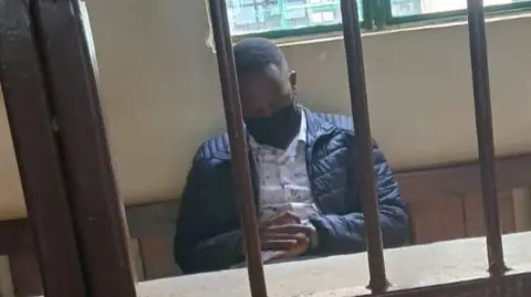 Kenya's Office of The Director of Prosecutions Festus Arasa Omwamba sits behind bars with his hands clasped, wearing a black face mask