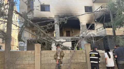 BBC A soldier in camouflage uniform and four other people - two wearing black high-viz jackets and hats, and a woman with long hair and a white shirt and a man with a dark grey T-shirt look over a surrounding wall into what appears to be the gutted section of a building that has been damaged