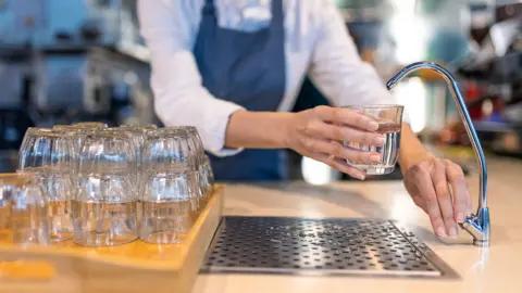Getty Images Stock photo shows a person filling a glass of water from a filter tap in a hospitality setting - they are positioned behind a bar with a stack of glasses in frame and wear a blue apron.