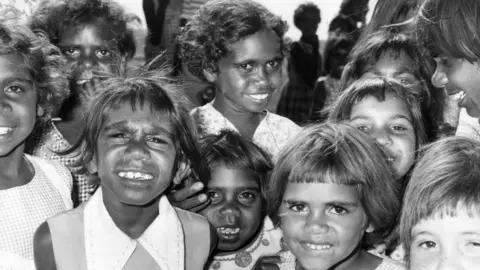 Getty Images A group of Aboriginal Australian children at Fitzroy Crossing in 1975. They're wearing checked dresses and smiling at the camera.