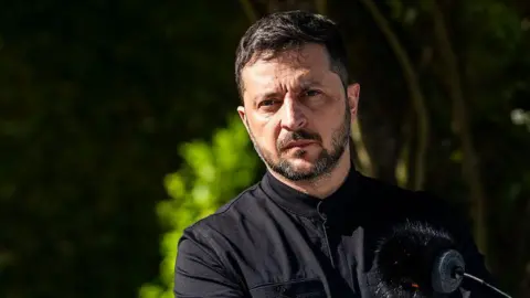 Getty Images A headshot of President Zelensky. He is wearing a black collarless shirt and standing outside, in front of a microphone. The background is blurry, but he appears to be standing in front of a tree or some foliage.