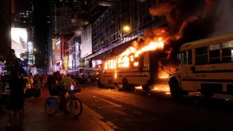 Reuters A yellow school bus is set alight with fire and smoke protruding from the vehicle at night time in Manhattan, New York City. Bystanders are gathered on the pavement opposite where the bus is parked, some taking videos on their phones. One bystander is sat on a bike.
