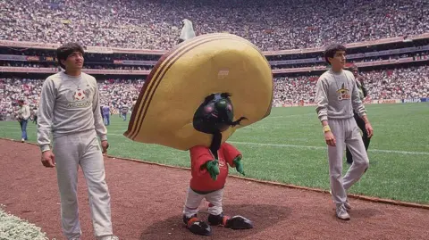 George Tiedemann/Sports Illustrated via Getty Images A small costumed figure wearing a red shirt and a large sombrero and large black-and-red shoes walks in the packed stadium between two men in Mexico 86 tracksuits.