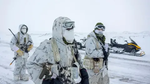 Reuters An image showing three Canadian military members in a snowy landscape, wearing white camouflage uniforms, face covers, gloves and holding various types of ammunition as they walk towards the right of the frame. Behind them is a parked snowmobile.