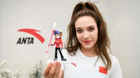 Getty Images Freestyle skier Eileen Gu posing for photos in front of the Anta logo on a wall, holding a mini figurine of herself