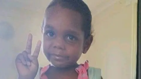 Northern Territory Police An Aboriginal girl wearing a pink dress and blue backpack poses for a picture, holding up the peace sign