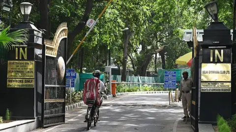 Getty Images A uniformed guard stands at the gate of the Delhi Gymkhana Club as a cyclist zooms into the campus surrounded by trees. Plates on the gates and the wall mention "Members only", "Rights of admission reserved", "No thorough fare", "Trespassers will be prosecuted", etc. 