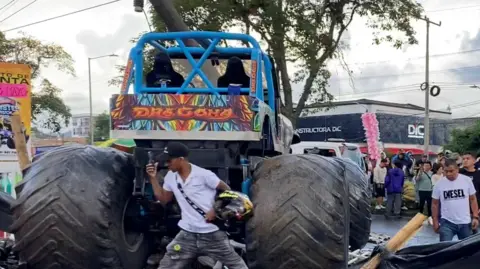 Reuters Image shows a blue monster truck which has come to halt after crashing through a barrier. It is surrounded by motorcycles that have fallen to the floor. People watch on. 
